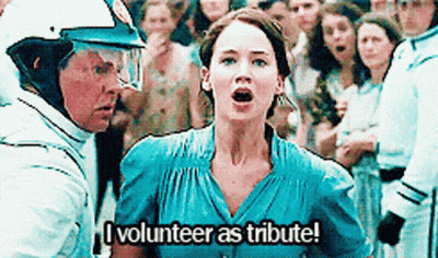 A Woman In A Blue Dress Is Standing In Front Of A Crowd Of People And Says `` I Volunteer As Tribute ! '' GIF