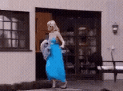 A Woman In A Blue Dress Is Standing In Front Of A House . GIF