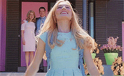 A Woman In A Blue Dress Is Standing In Front Of A Pink House With Her Arms Outstretched . GIF