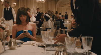 A Woman In A Blue Dress Sits At A Table Looking At Her Cell Phone GIF