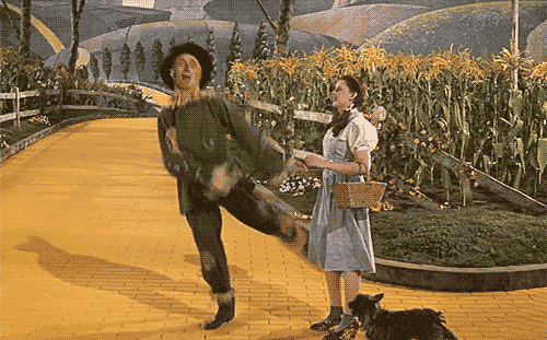 A Woman In A Blue Dress Stands Next To A Scarecrow On A Yellow Brick Road GIF