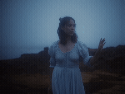 A Woman In A Blue Dress Stands On A Rocky Shore GIF