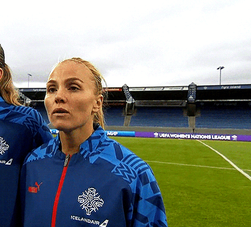 A Woman In A Blue Icelandair Jacket Stands In Front Of A Soccer Field GIF