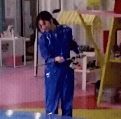 A Woman In A Blue Jumpsuit Is Standing In A Room With A Hose . GIF