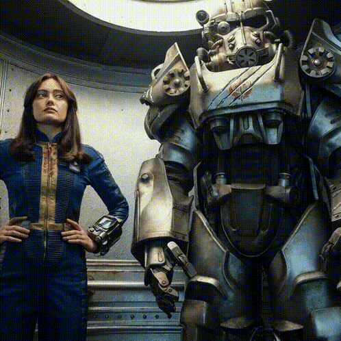 A Woman In A Blue Jumpsuit Stands Next To A Robot With A Red X On Its Chest GIF