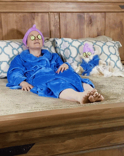 A Woman In A Blue Robe Is Laying On A Bed Next To A Small White Dog GIF