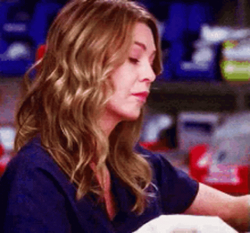 A Woman In A Blue Scrub Top Is Sitting At A Table And Looking At Something . GIF