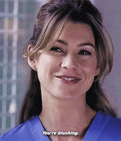 A Woman In A Blue Scrub Top Is Smiling And Saying You 'Re Blushing . GIF