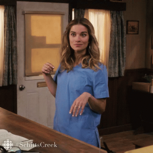 A Woman In A Blue Scrub Top Is Standing In Front Of A Door With #Schittscreek Written On It GIF