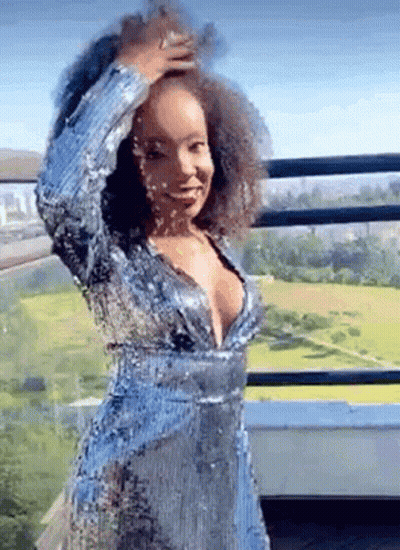 A Woman In A Blue Sequined Dress Is Standing On A Balcony Holding Her Hair . GIF