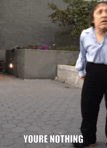 A Woman In A Blue Shirt And Black Pants Is Standing On A Brick Sidewalk With The Words " Youre Nothing " Above Her GIF