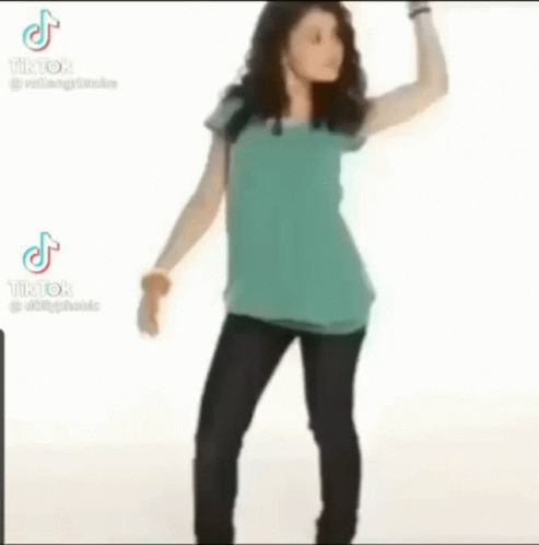 A Woman In A Blue Shirt And Black Pants Is Standing With Her Hands On Her Hips And Looking At The Camera . GIF