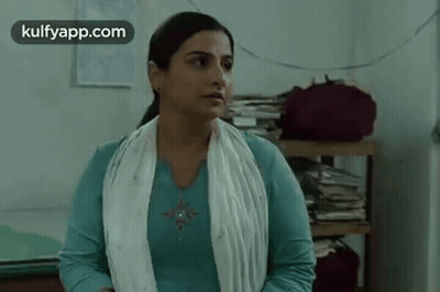 A Woman In A Blue Shirt And White Scarf Is Standing In A Room With A Stack Of Books On A Shelf . GIF