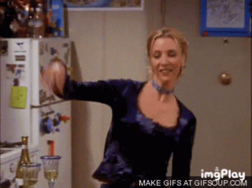 A Woman In A Blue Shirt Is Dancing In Front Of A Refrigerator And A Bottle Of Wine GIF