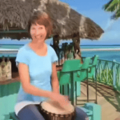 A Woman In A Blue Shirt Is Playing A Drum On The Beach GIF