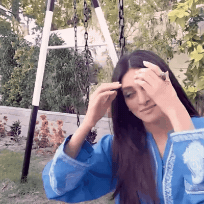 A Woman In A Blue Shirt Is Sitting On A Swing Adjusting Her Hair GIF