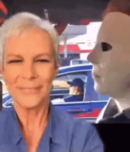 A Woman In A Blue Shirt Is Smiling In Front Of A Man In A Mask . GIF