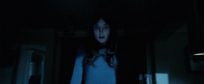 A Woman In A Blue Shirt Is Standing In A Dark Room Looking At The Camera GIF