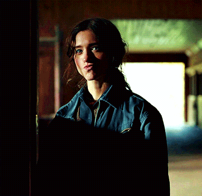 A Woman In A Blue Shirt Is Standing In A Dark Room Looking At The Camera GIF