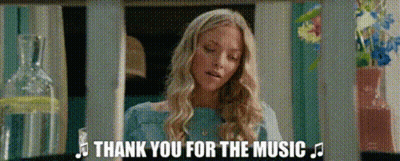 A Woman In A Blue Shirt Is Standing In Front Of A Bottle Of Water And Flowers And Says Thank You For The Music . GIF