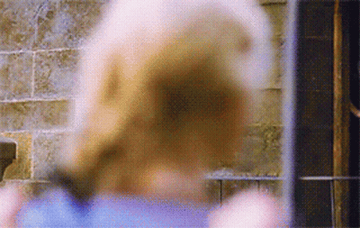 A Woman In A Blue Shirt Is Standing In Front Of A Brick Wall Looking Out A Window . GIF