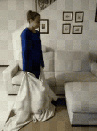 A Woman In A Blue Shirt Is Standing In Front Of A White Couch Holding A White Sheet GIF