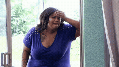 A Woman In A Blue Shirt Is Standing In Front Of A Window And Covering Her Forehead With Her Hand . GIF