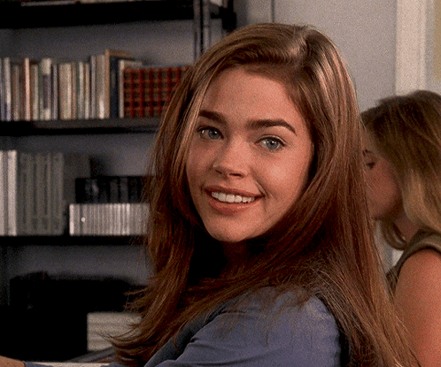 A Woman In A Blue Shirt Smiles In Front Of A Bookshelf GIF