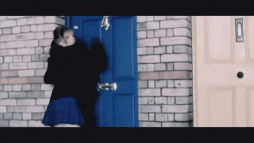 A Woman In A Blue Skirt Stands In Front Of A Blue Door With A Brick Wall Behind Her GIF