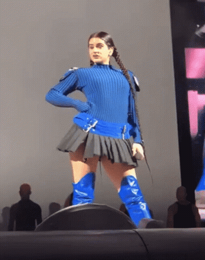 A Woman In A Blue Sweater And Blue Boots Stands On A Stage Holding A Microphone GIF
