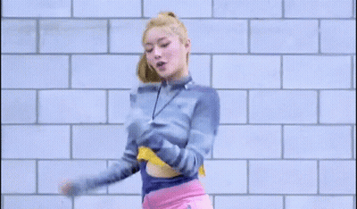 A Woman In A Blue Sweater And Pink Shorts Is Dancing In Front Of A Brick Wall . GIF