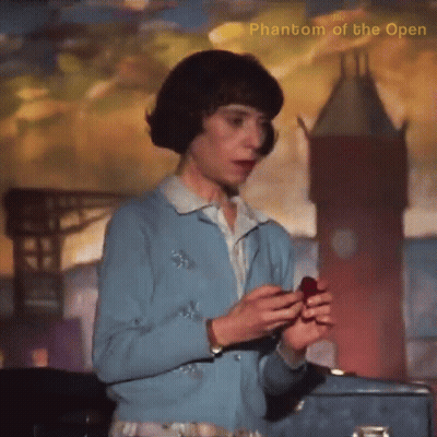 A Woman In A Blue Sweater Is Holding A Red Object In Front Of A Phantom Of The Open Painting GIF