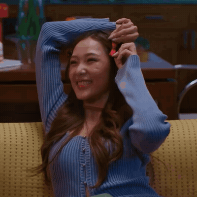 A Woman In A Blue Sweater Is Sitting On A Yellow Couch And Smiling GIF