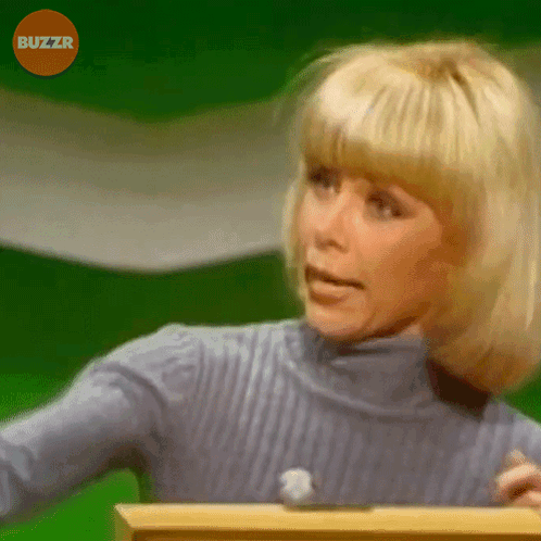 A Woman In A Blue Sweater Is Standing In Front Of A Green Screen With Buzzr Written On It GIF