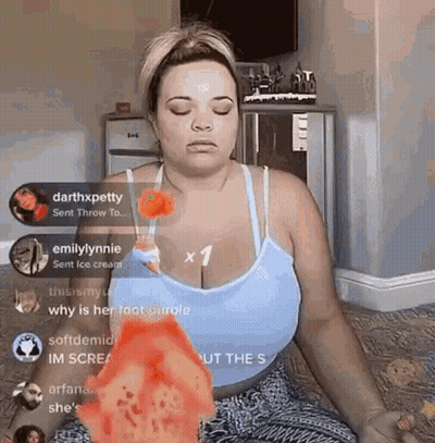 A Woman In A Blue Tank Top Is Sitting On The Floor With Her Eyes Closed And Holding A Piece Of Pizza . GIF
