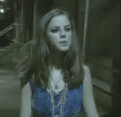 A Woman In A Blue Top And Necklace Is Standing In A Dark Room . GIF