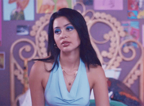 A Woman In A Blue Top Is Sitting On A Bed With A Purple Headboard GIF