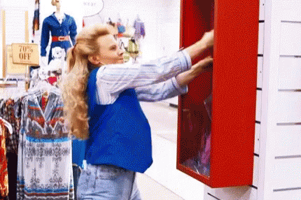 A Woman In A Blue Vest Is Reaching Into A Red Closet In A Store . GIF