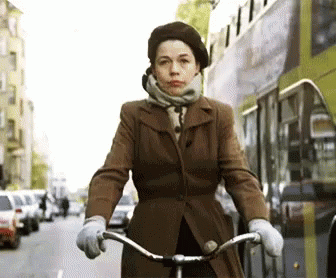 A Woman In A Brown Coat Is Riding A Bicycle Down A City Street GIF