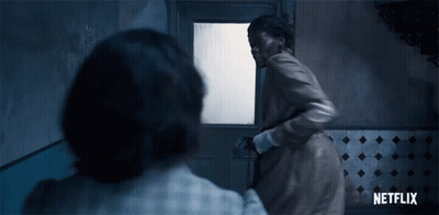 A Woman In A Brown Coat Is Standing In Front Of A Door In A Room . GIF