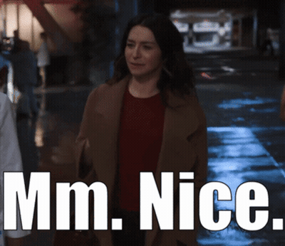 A Woman In A Brown Coat Is Walking Down A Street With The Words Mm Nice Behind Her GIF