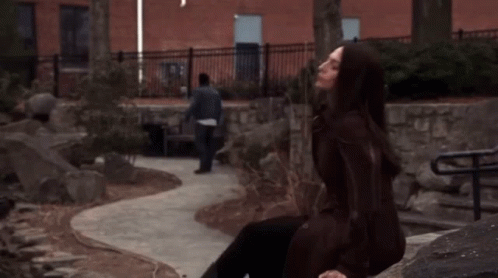 A Woman In A Brown Coat Sits On A Rock While A Man Walks By GIF
