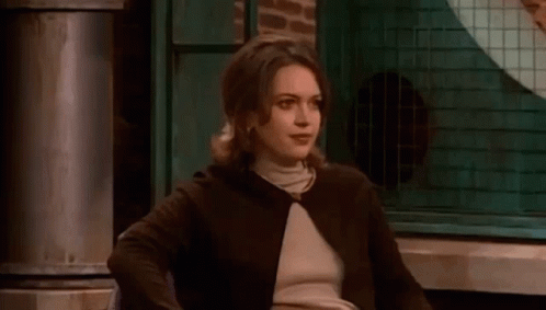 A Woman In A Brown Jacket And Turtleneck Is Sitting In A Chair In Front Of A Window . GIF
