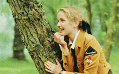 A Woman In A Brown Jacket Is Leaning Against A Tree Trunk And Smiling GIF