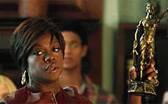 A Woman In A Brown Jacket Is Standing In Front Of A Statue Of A Woman . GIF