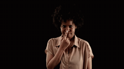 A Woman In A Brown Shirt Holds Her Finger To Her Nose Against A Black Background GIF
