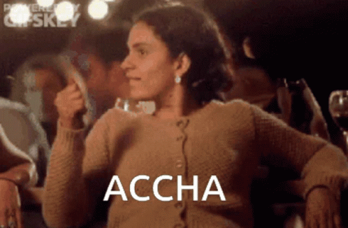 A Woman In A Brown Sweater Is Sitting In Front Of A Group Of People And The Word Accha Is On The Screen GIF
