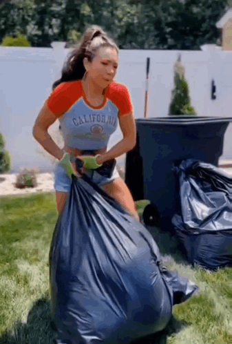 A Woman In A California Shirt Is Holding A Large Garbage Bag . GIF