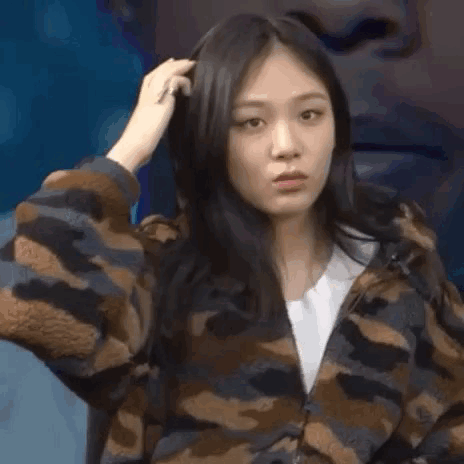 A Woman In A Camouflage Jacket Is Looking At The Camera While Holding Her Hair . GIF