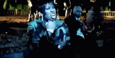 A Woman In A Camouflage Jacket Sings Into A Microphone In Front Of A Man In A Suit GIF